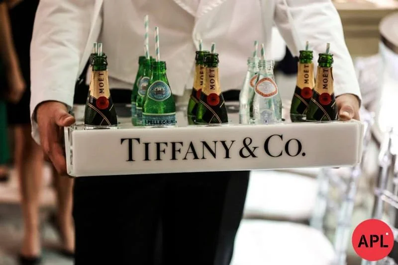 Waiter holding a tray with small bottles of Moët champagne and sparkling water, with a Tiffany & Co. logo on the tray, at an event.