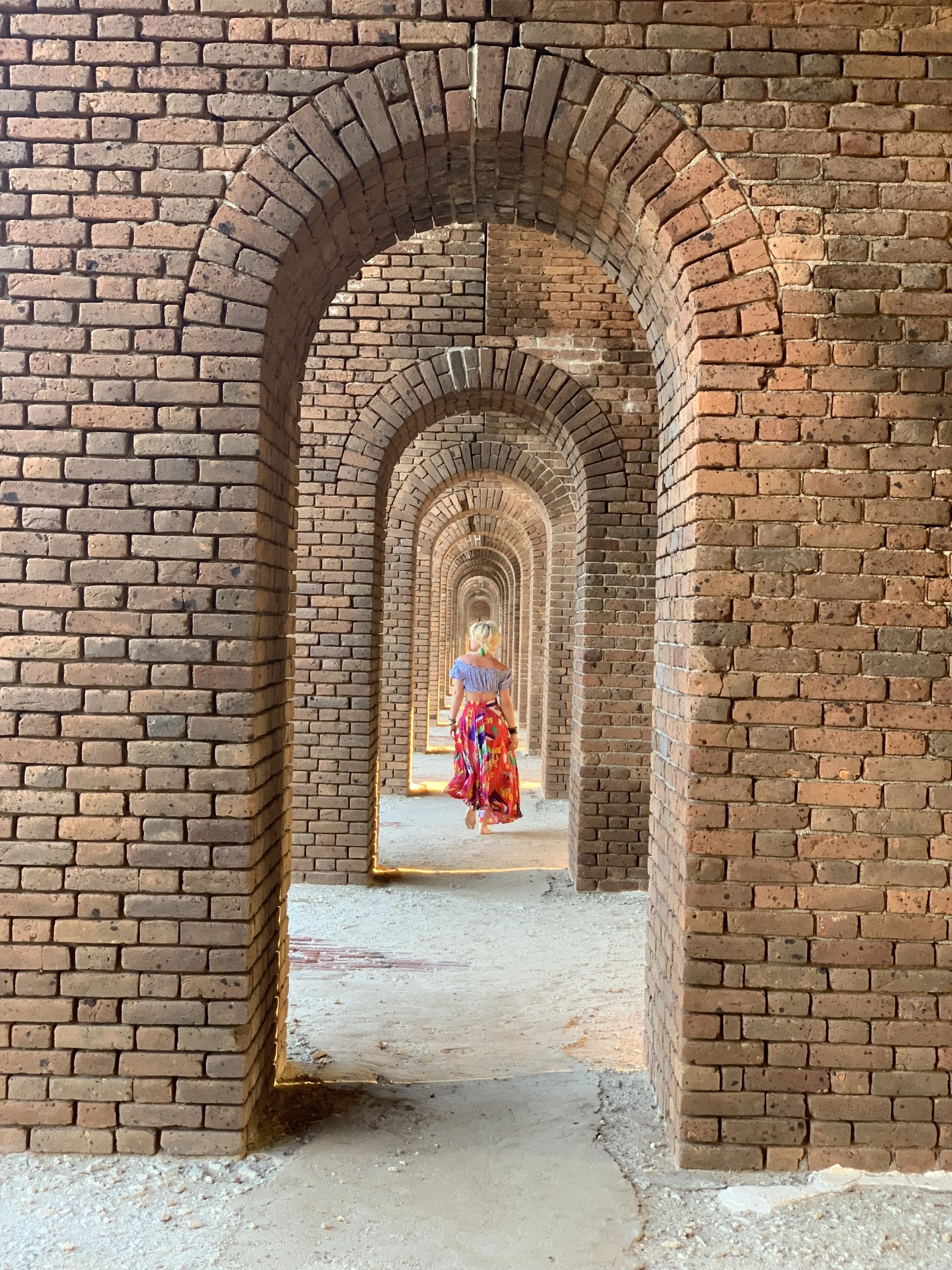 Photo of brick arches built in succession with a woman in a flowing pink skirt and blue cropped blouse walking away from the camera, through the arches, looking into the sun.