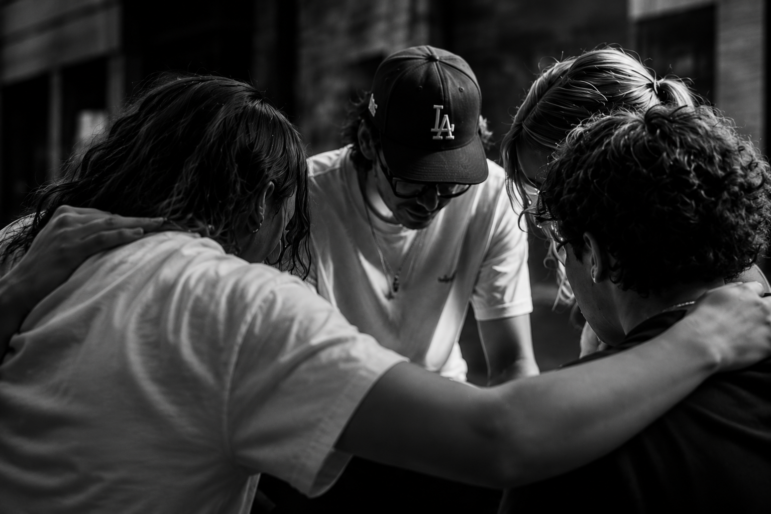 Group of five people, standing in a circle with heads bowed, engaging in a prayer or moment of reflection indoors.
