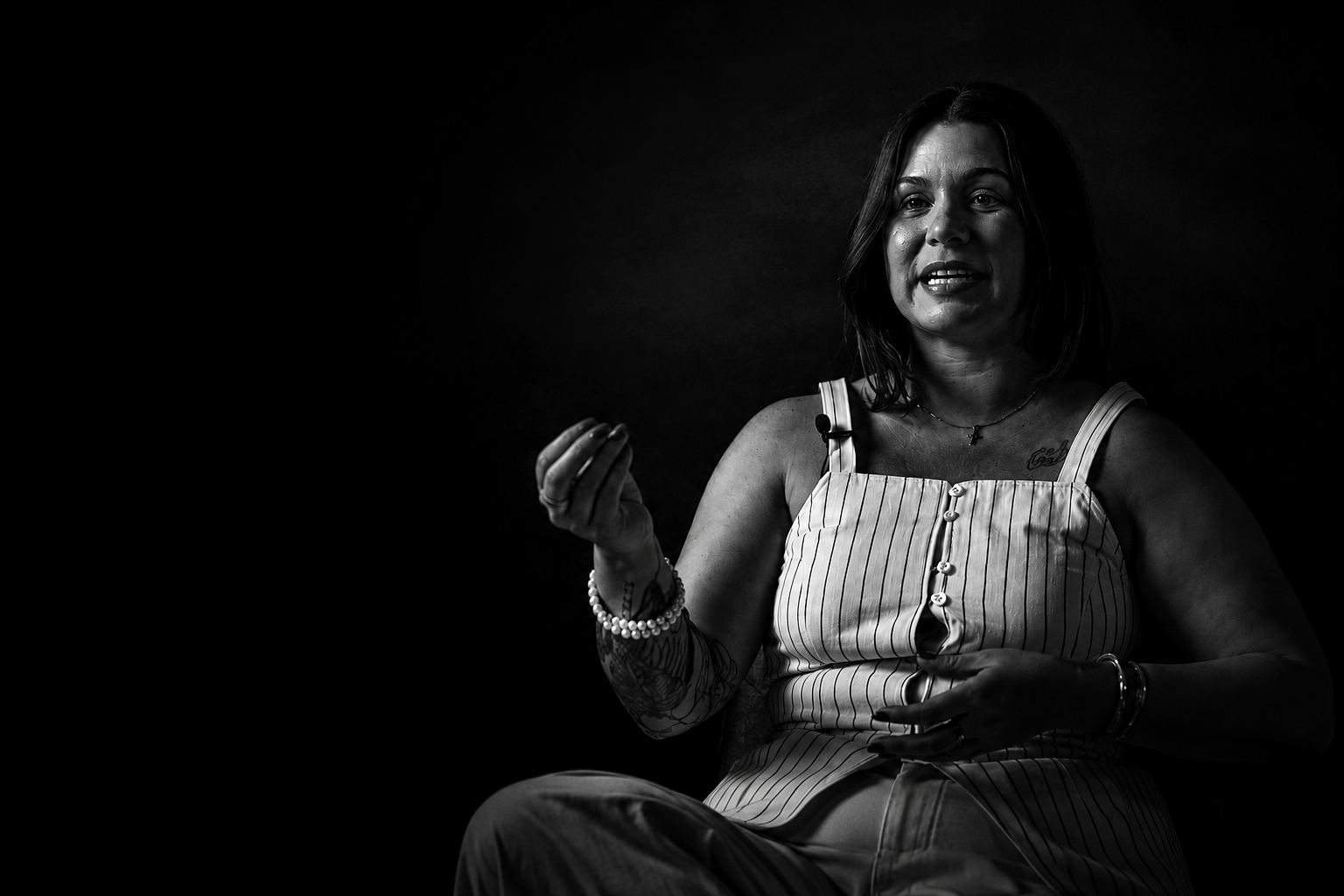 A woman with shoulder-length dark hair, wearing a striped sleeveless top, sits against a dark background, gesturing with her right hand while speaking.