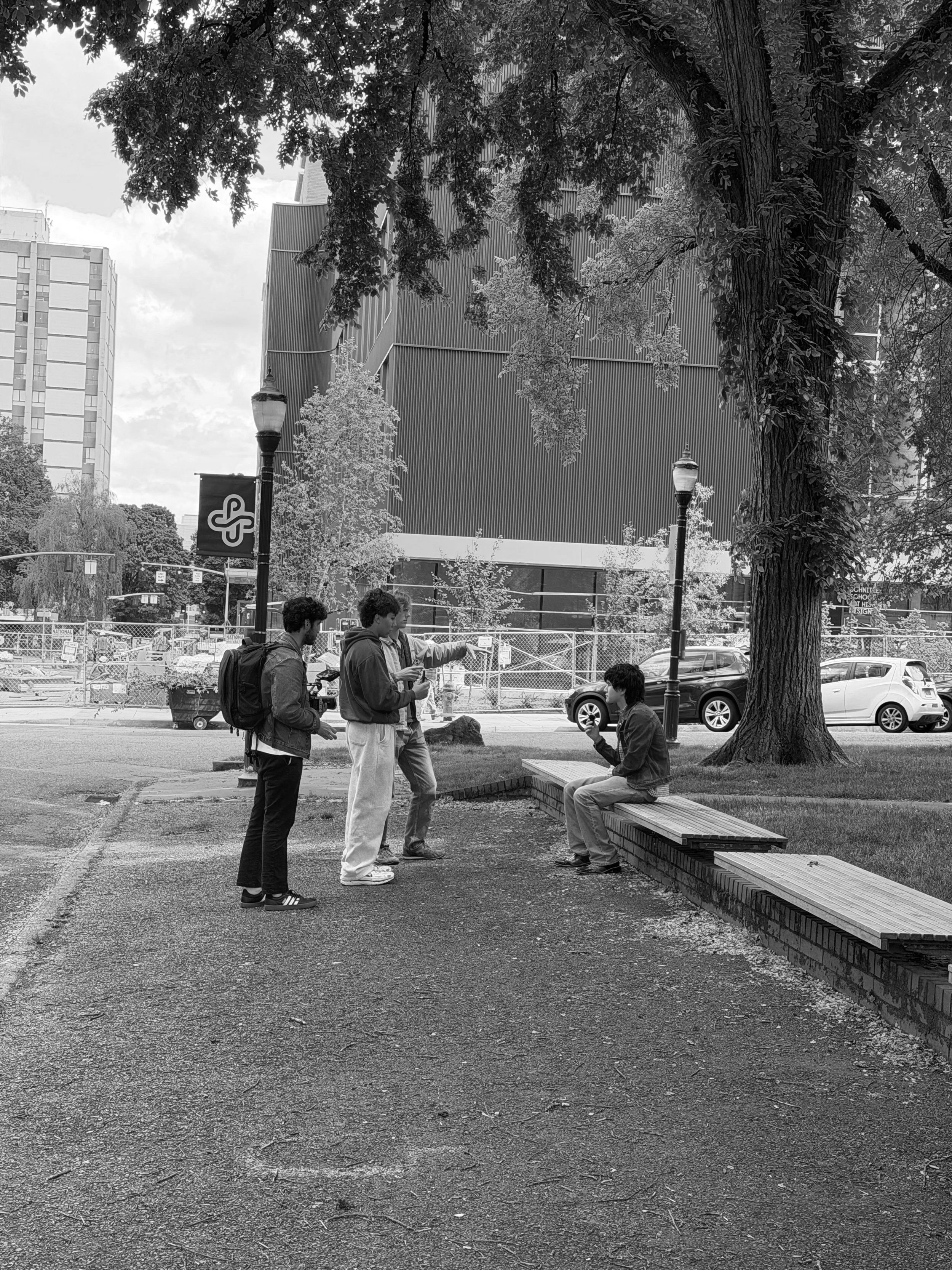 Four young males, three standing and one sitting on a wooden bench, are in conversation outdoors near a large tree with textured bark. In the background, there are buildings, a street with cars parked, and street lamps. The scene is in black and white.