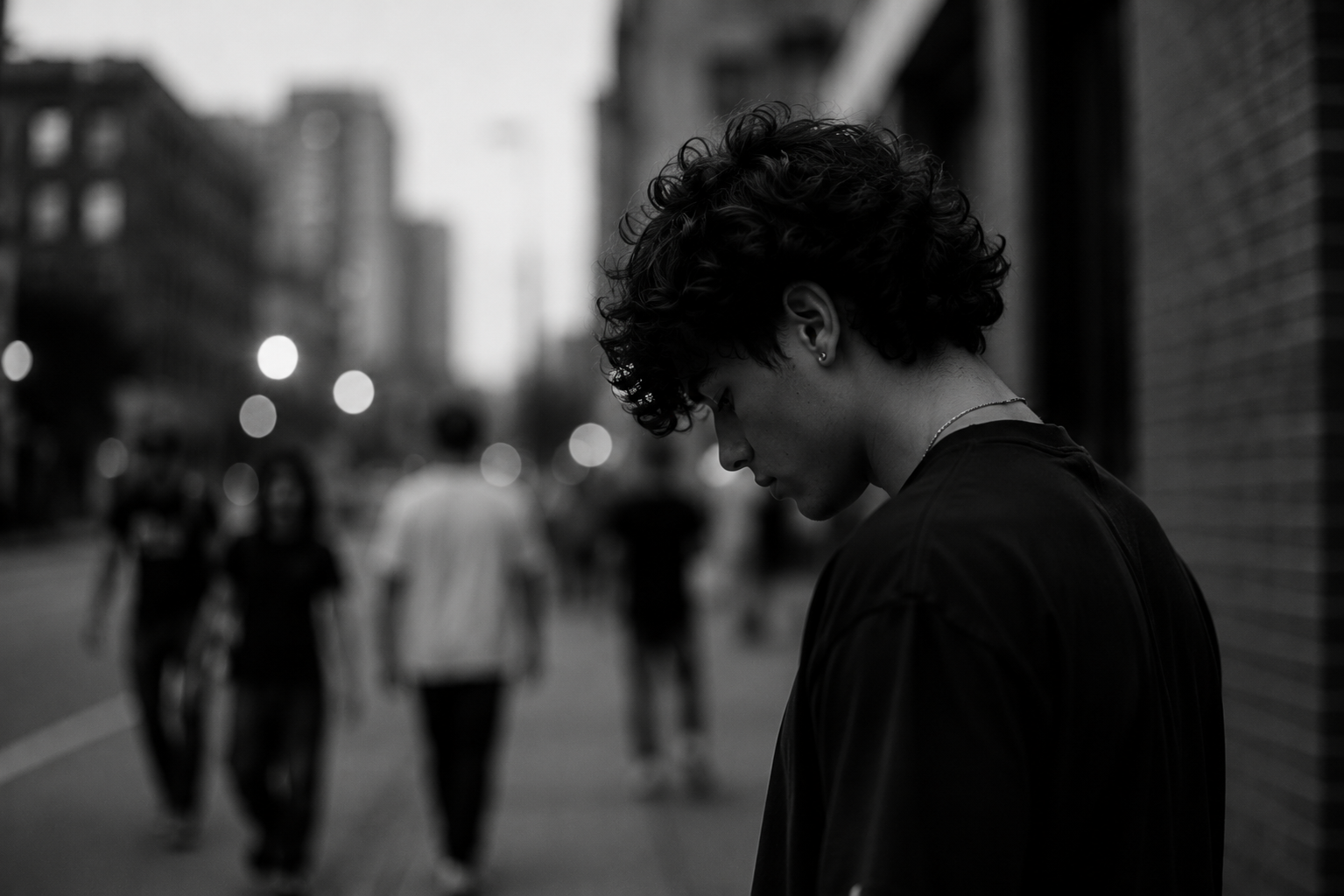 A black and white photo of a young man with curly hair looking down on a city street at dusk, with blurred pedestrians in the background.