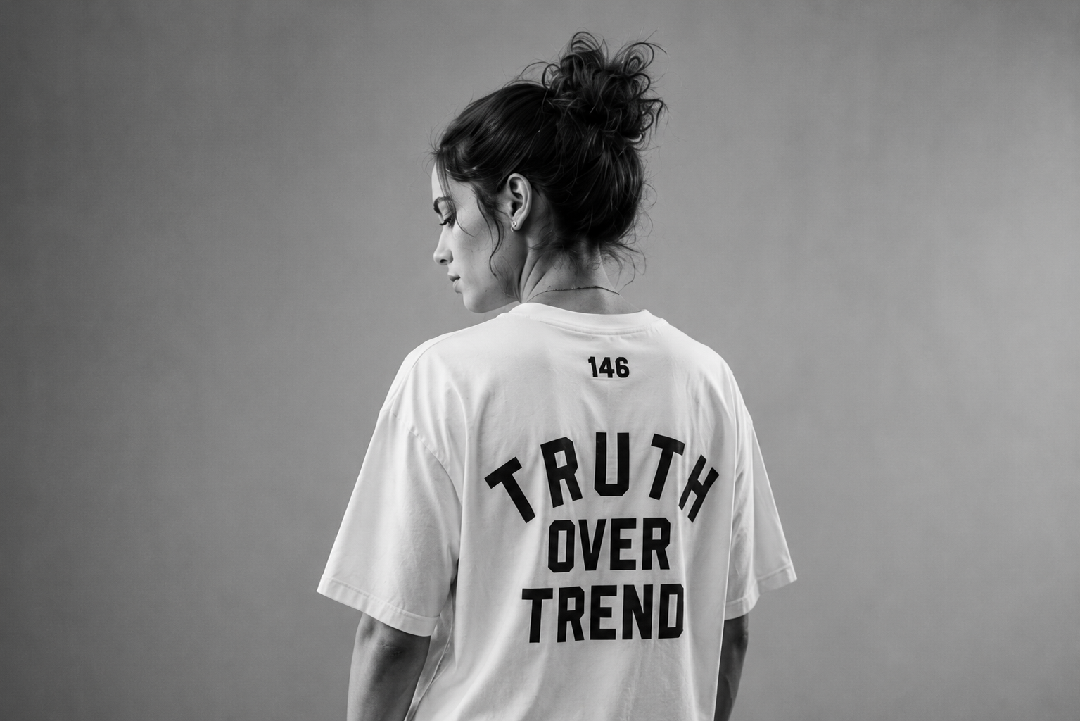 A woman with curly hair tied in a bun, in profile, wearing a loose white T-shirt with the words "TRUTH OVER TREND" printed on the back, standing against a plain gray background.