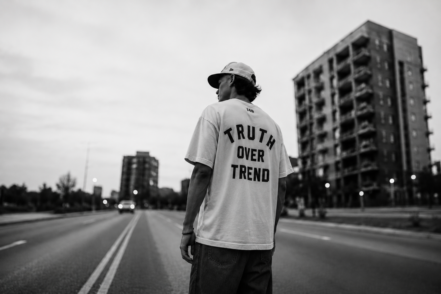 A person standing in the middle of a city street wearing a white t-shirt that says 'TRUTH OVER TREND' on the back, with tall buildings in the background, in black and white.
