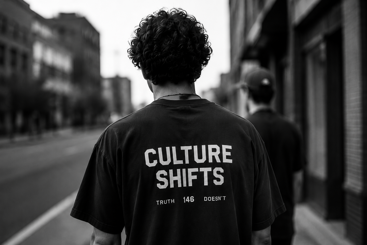 Back view of a person with curly hair wearing a T-shirt that says 'CULTURE SHIFTS' and 'TRUTH 146 DOESN'T' walking down an urban sidewalk.