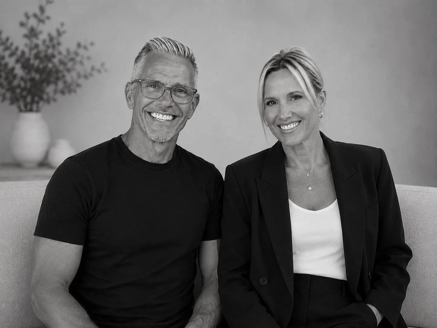 Black and white photo of a smiling man with glasses and a woman in business attire sitting on a couch in a room with a vase and decorative objects in the background.