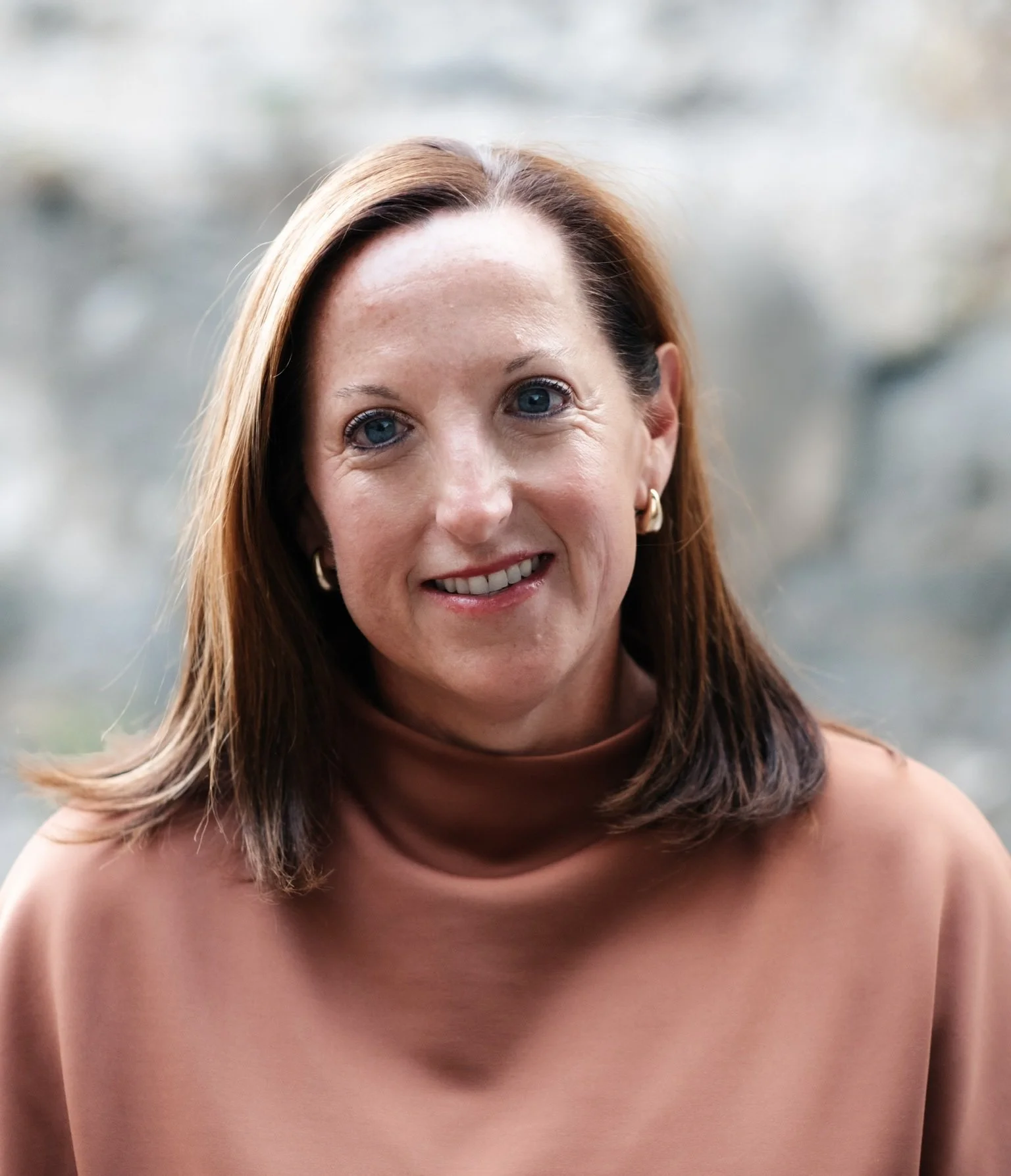 Close-up image of a smiling woman with red hair, blue eyes, gold earrings, and a tan shirtless upper body.