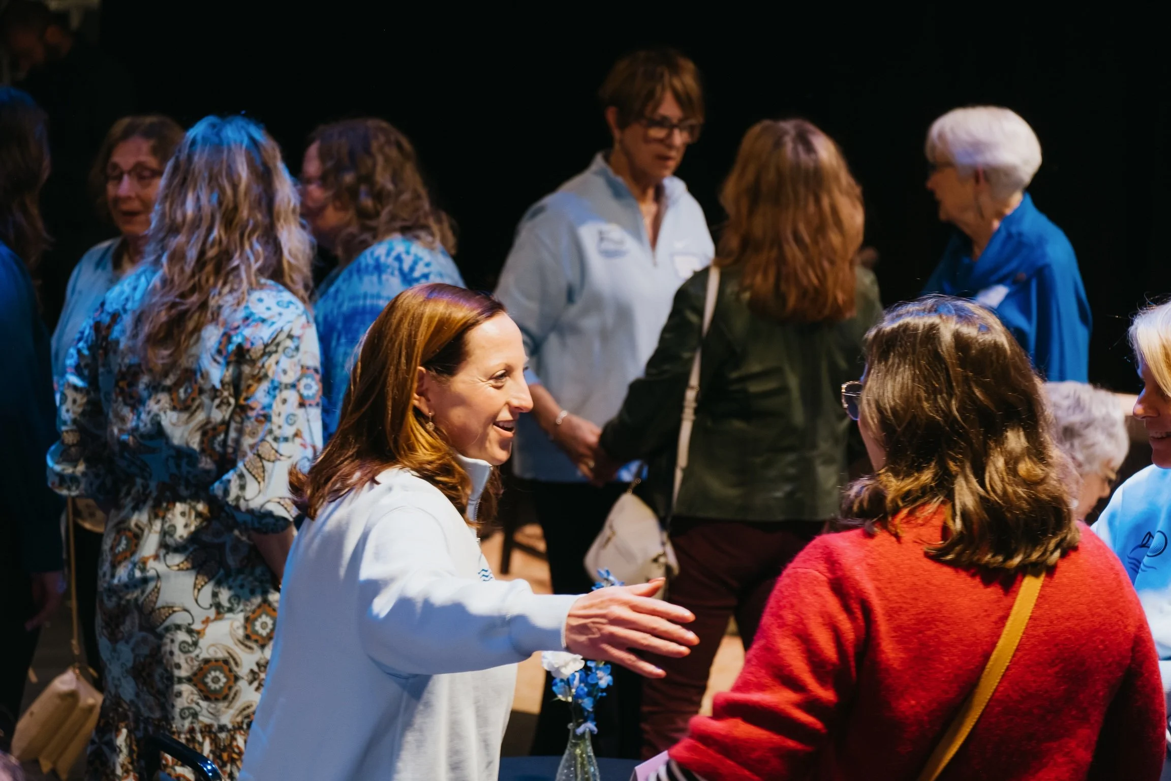 A group of women engaged in conversation, including one woman with red hair smiling and another woman with gray hair in a blue jacket, in a dark indoor setting.