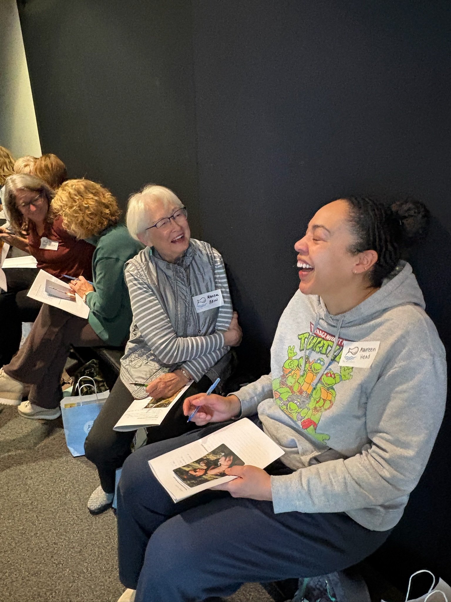A group of women sitting in a line, smiling and engaging in conversation. They are holding papers and pens, and appear to be at a workshop or seminar. The woman in front is wearing a Teenage Mutant Ninja Turtles hoodie, and the second woman has a nam