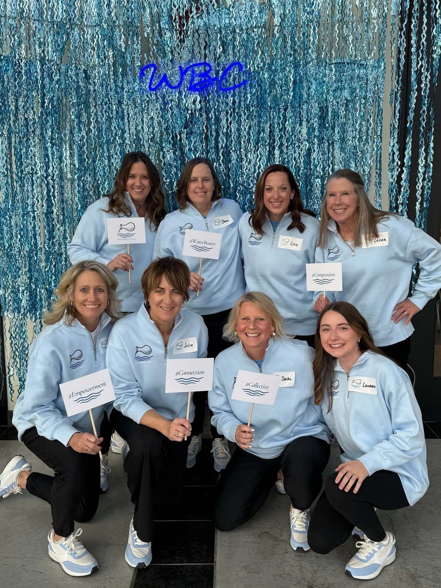 Group of six women posing in front of a blue sequin curtain. They are wearing matching light blue jackets with water-related logos and carrying signs with various hashtags and words like '#WaterBearer,' '#Empowerment,' 'Connection,' '#Collective,' an