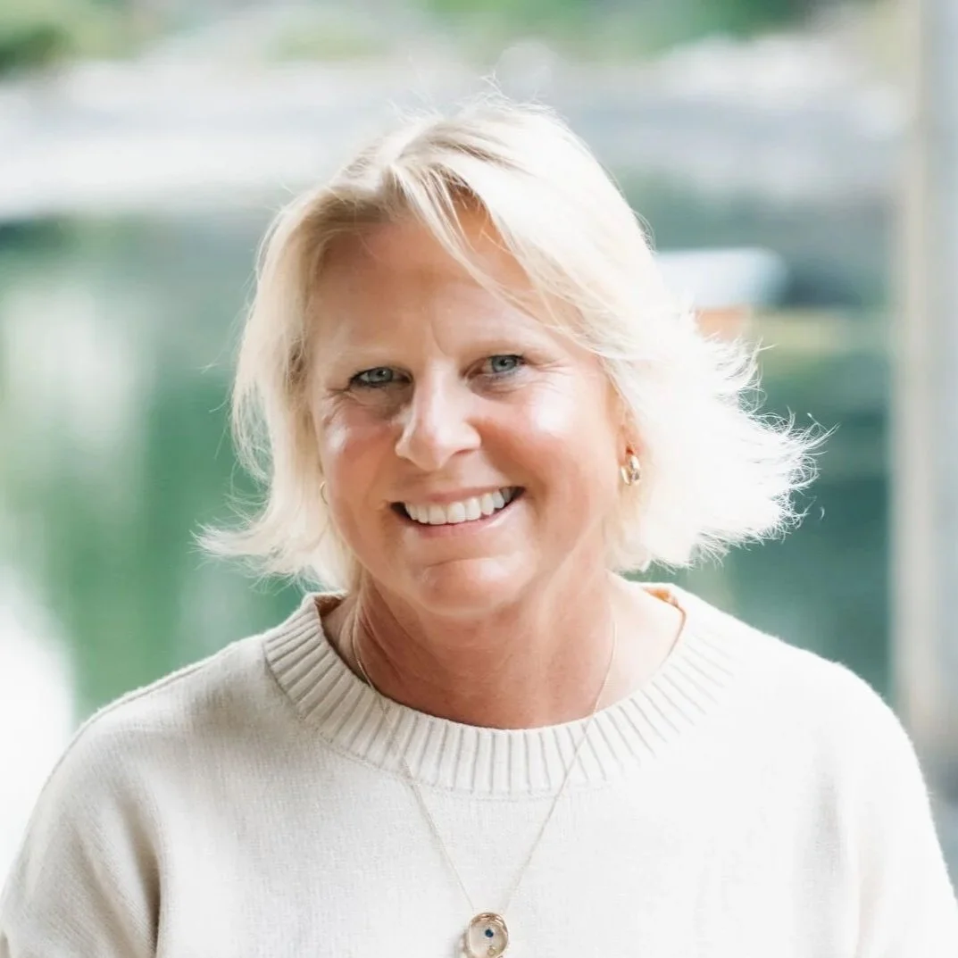 A smiling woman with short blonde hair, wearing a cream sweater and a necklace with a round pendant, in front of a blurred background.