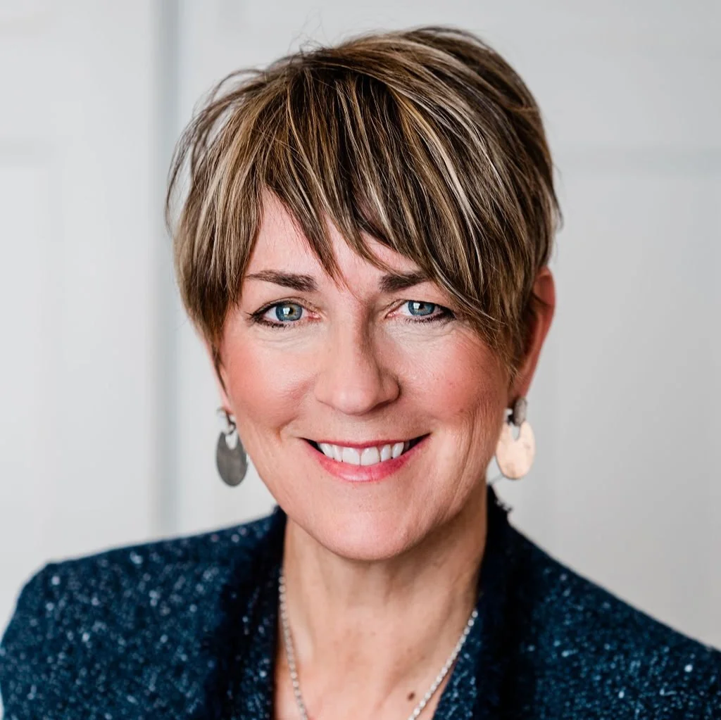 Close-up of a woman with short brown hair and blue eyes, smiling, wearing earrings and a dark blue blazer.