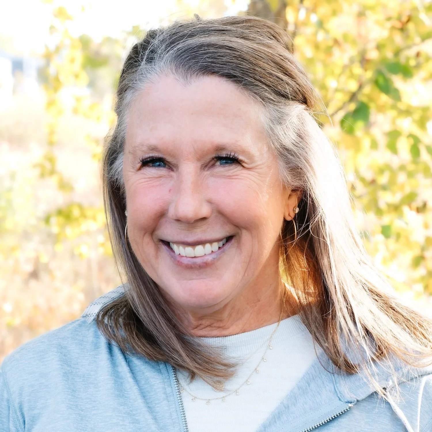 A middle-aged woman with gray hair and blue eyes smiling outdoors, with trees and sunlight in the background.