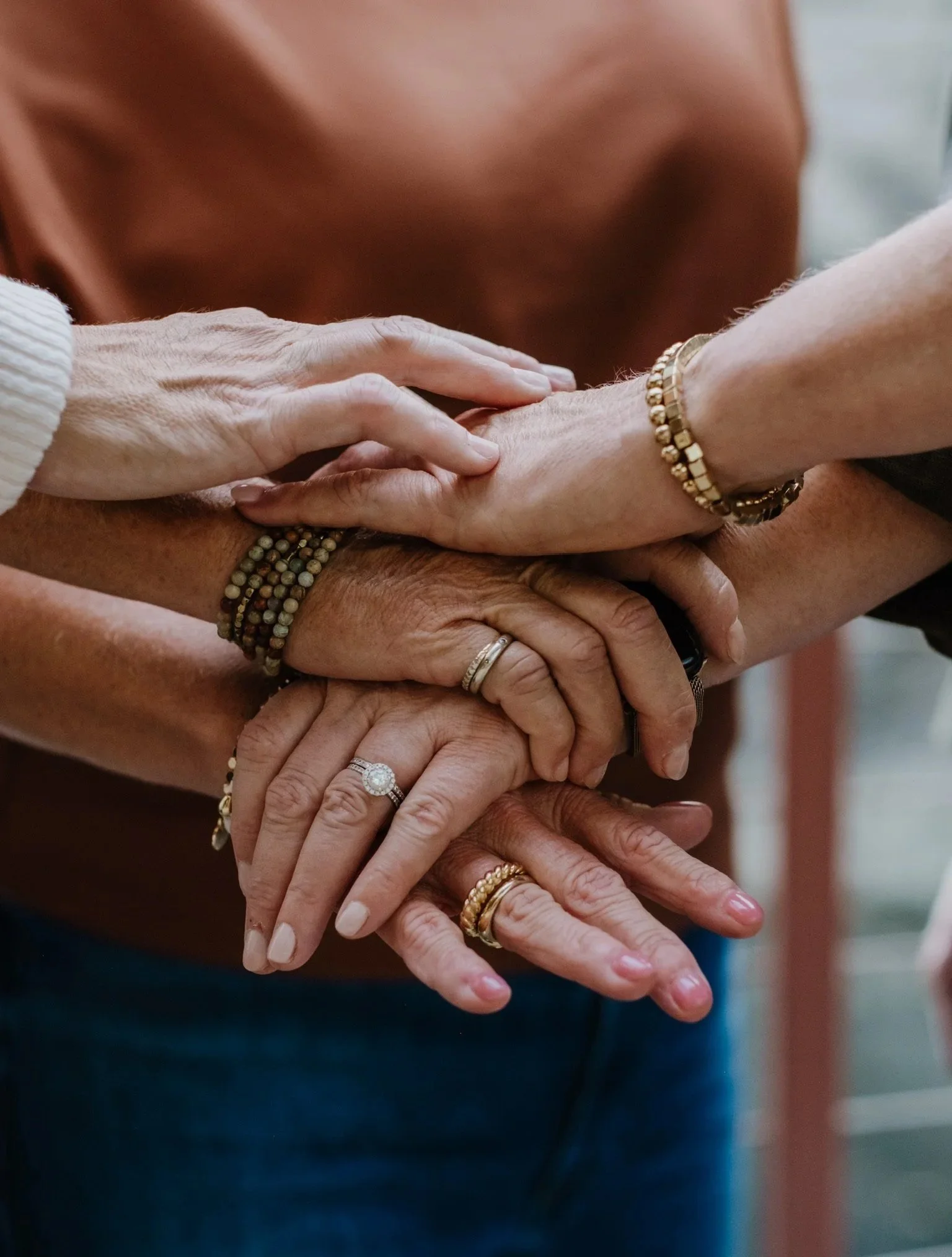 Multiple hands stacked together, showing diverse skin tones and jewelry, symbolizing unity and teamwork.