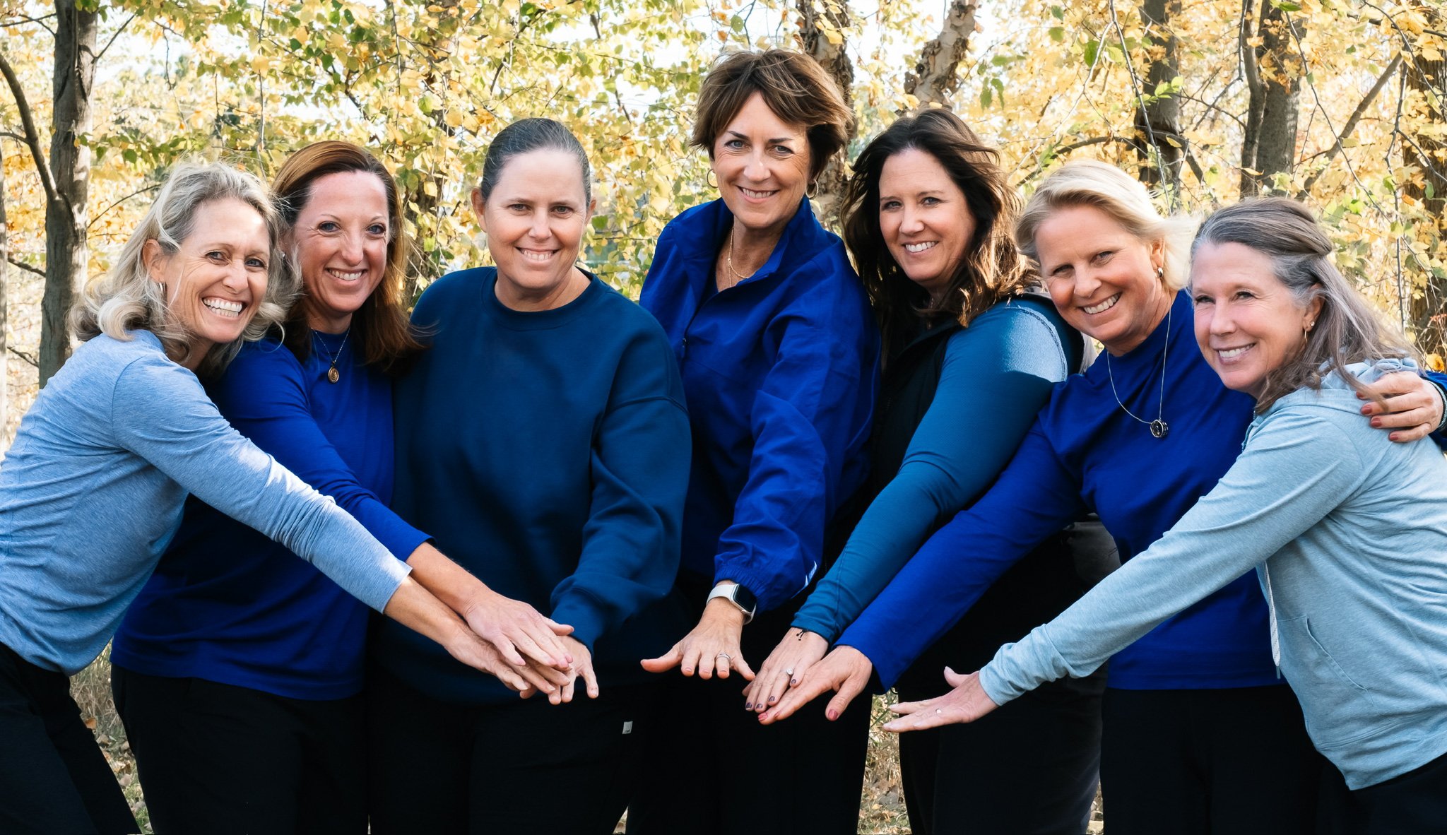 Seven women, the founders of Water Bearer Collective, standing outdoors in front of trees with yellow leaves, smiling, with their hands stacked together in a sign of unity.