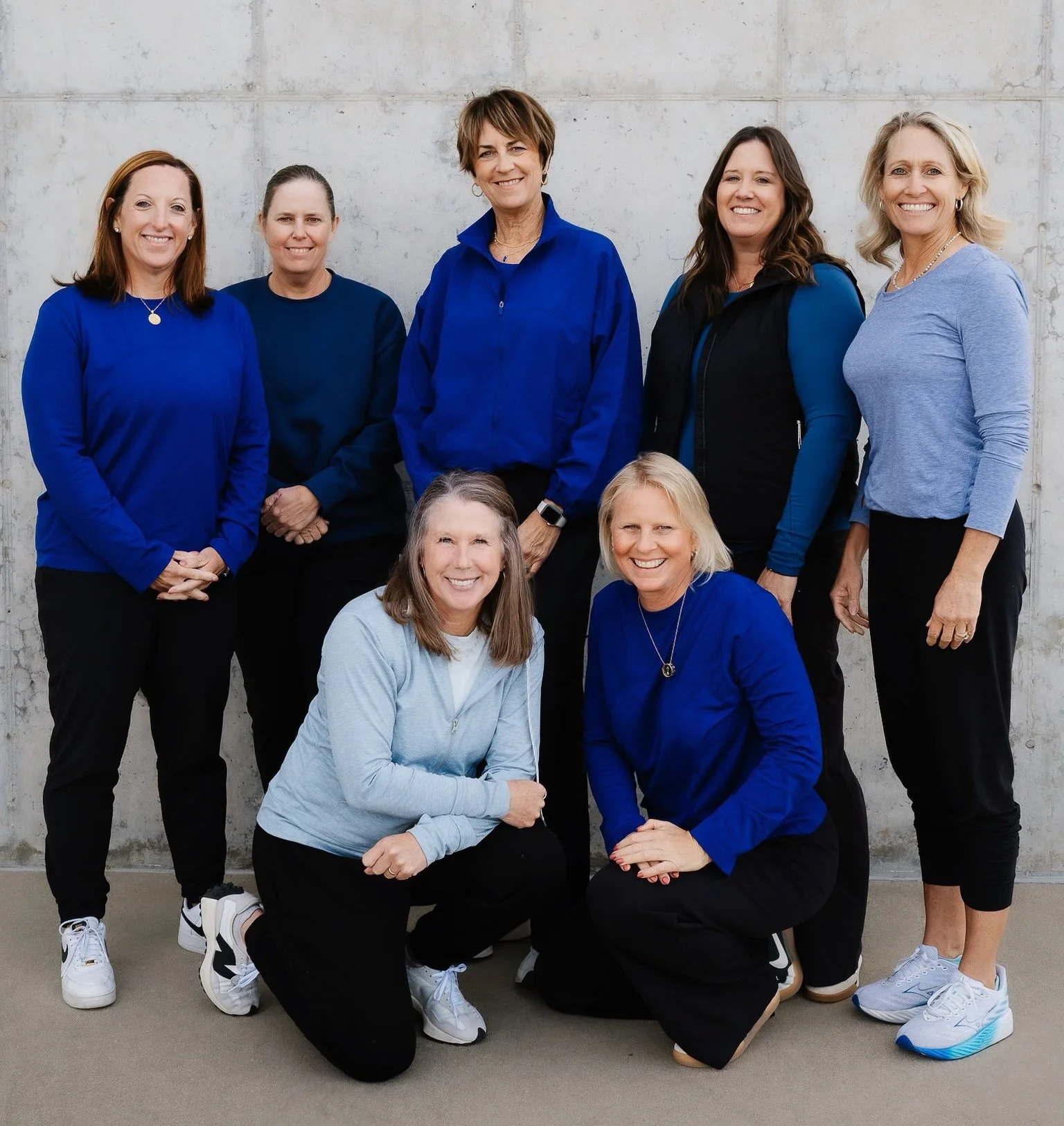 Group of nine women, the founders of Water Bearer Collective in Iowa City, posing together against a concrete wall, some standing and some kneeling, all smiling and wearing casual clothing in shades of blue.