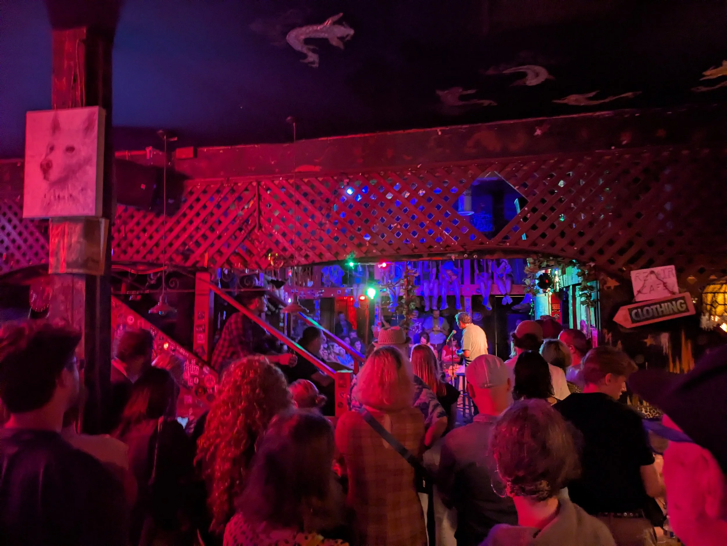 A crowded indoor bar or club with red and purple lighting, showing many people gathered around. The ceiling is decorated with hanging palm trees, and there are various signs and decorations on the walls.