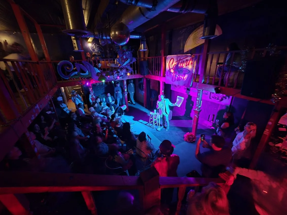 A  is taking place in a dimly lit venue with neon signsThe stage is set with a performer holding a microphone, and the audience is seated in front of the stage, watching the performance. The venue has a cozy, club-like atmosphere with colorful lighting.