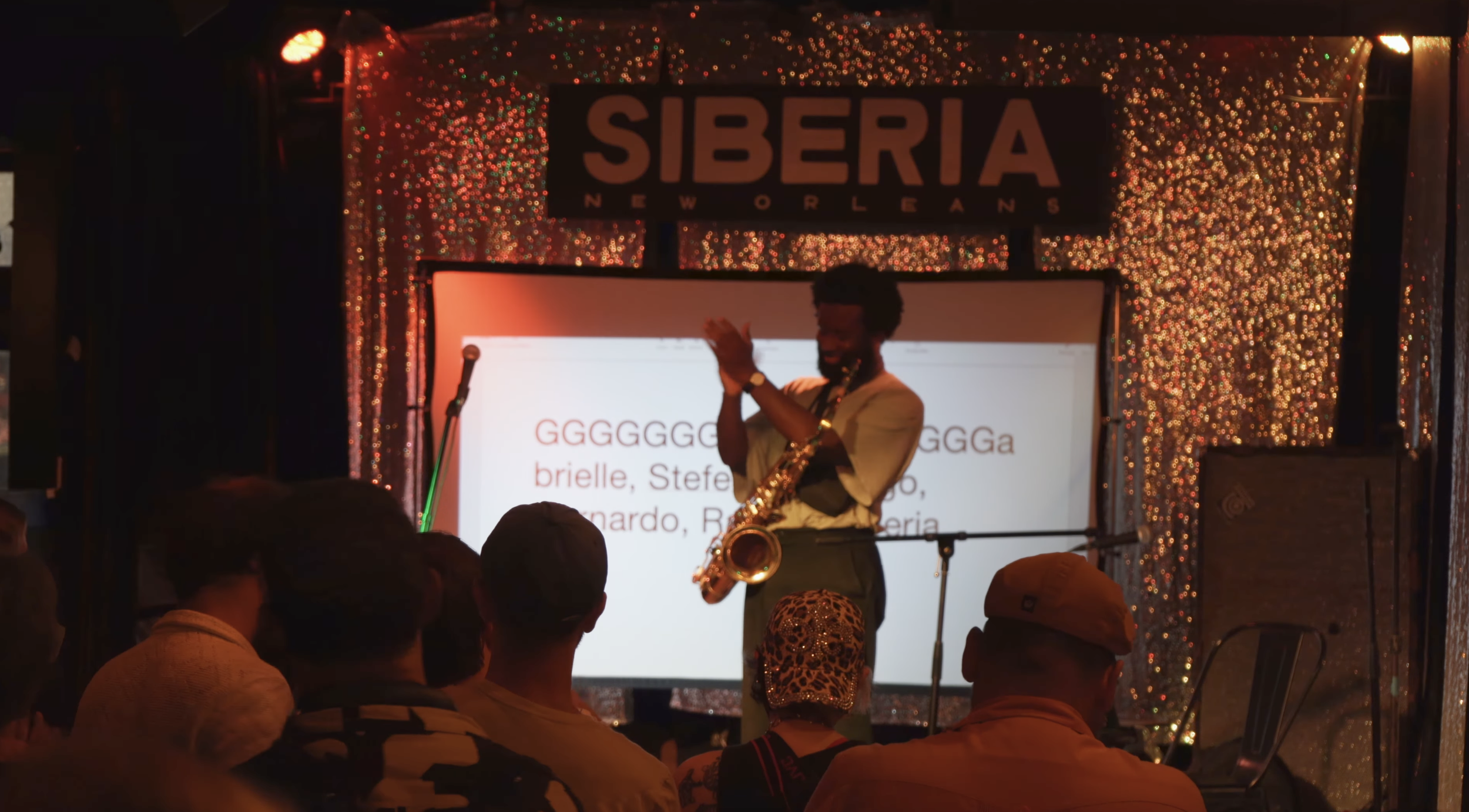 A man playing a saxophone on stage at a Siberia New Orleans event, with a large sign reading 'SIBERIA NEW ORLEANS' behind him. The stage is decorated with multicolored string lights. Audience members are seated, watching the performance.