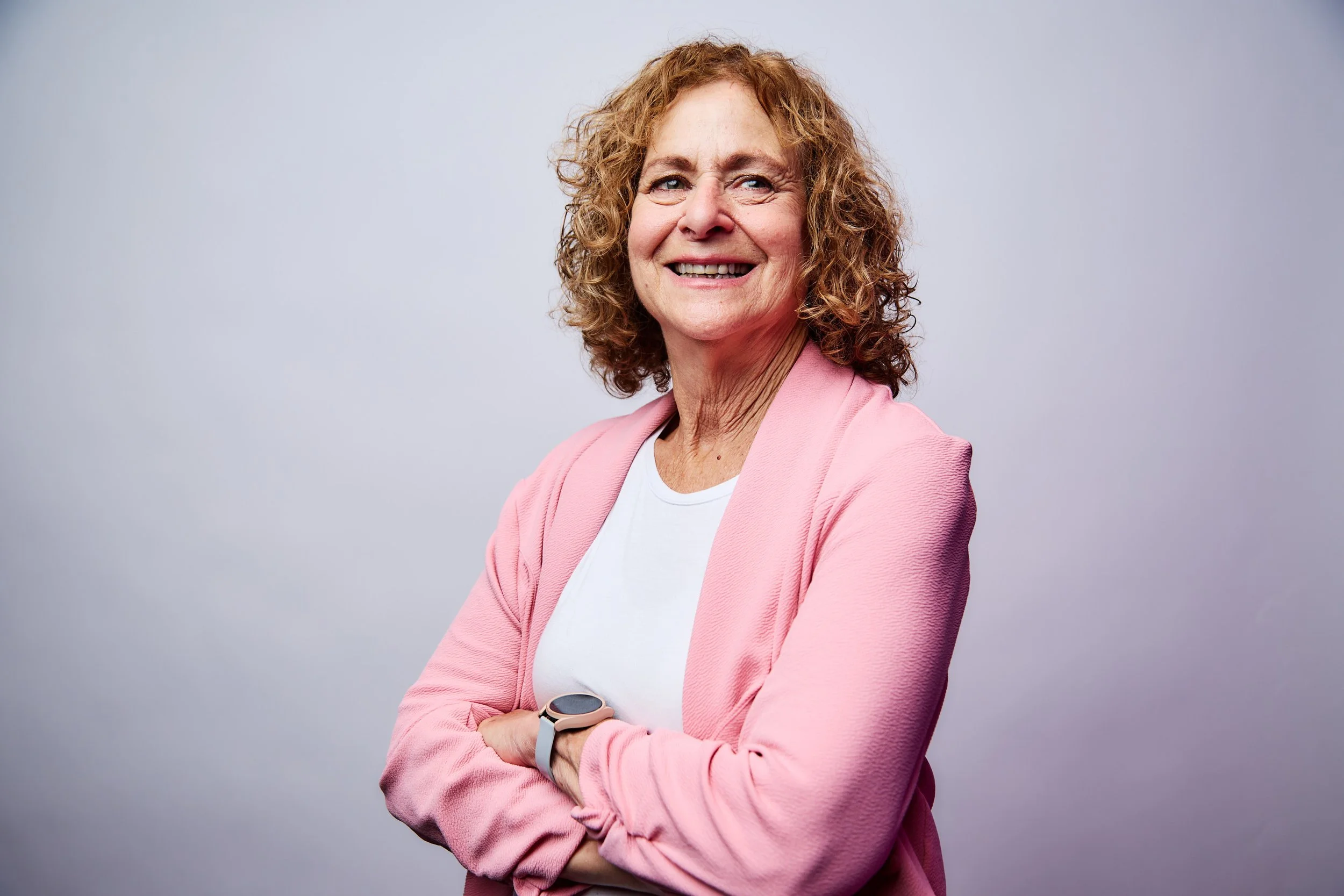A smiling woman with curly red hair wearing a pink blazer over a white shirt, standing against a neutral background.