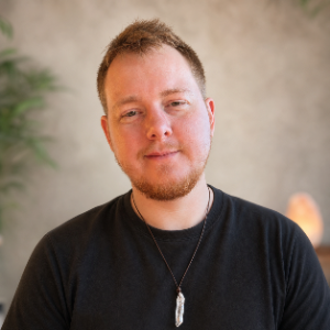 A young man with short light brown hair, a beard, wearing a black shirt and necklace, smiling against a neutral background.