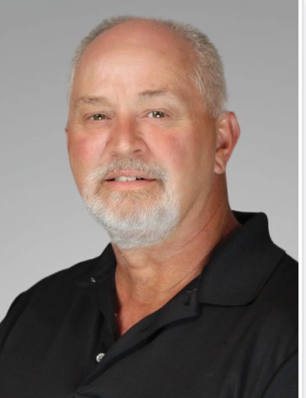 Headshot of a middle-aged man with short gray hair and a beard, wearing a black polo shirt, against a plain gray background.