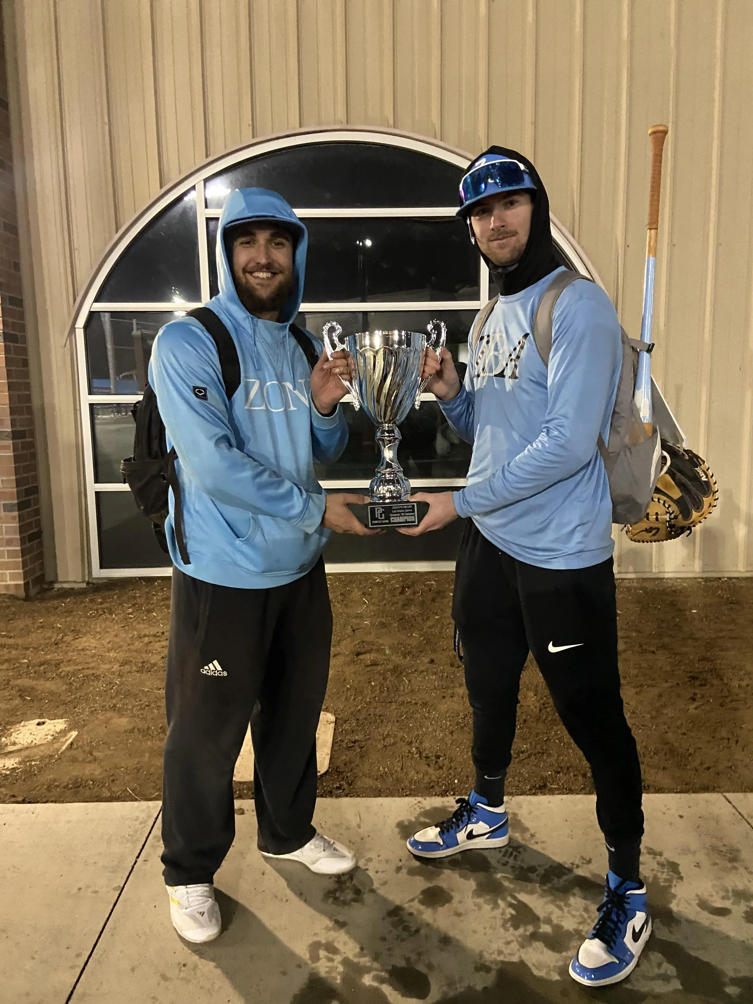 Two young men in athletic clothing and backpacks standing outside at night, holding a large trophy together. One wears a light blue hoodie, black pants, and white sneakers; the other wears a light blue long-sleeve shirt, black pants, and blue and white sneakers. They are smiling and standing in front of a building with a semi-circle window.
