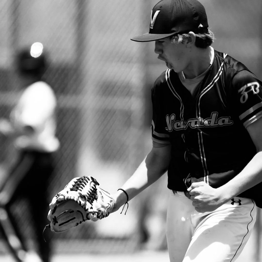 A male baseball player wearing a cap, jersey with 'Nevada' on it, and white pants, appears to be shouting or yelling while holding a baseball glove during a game.
