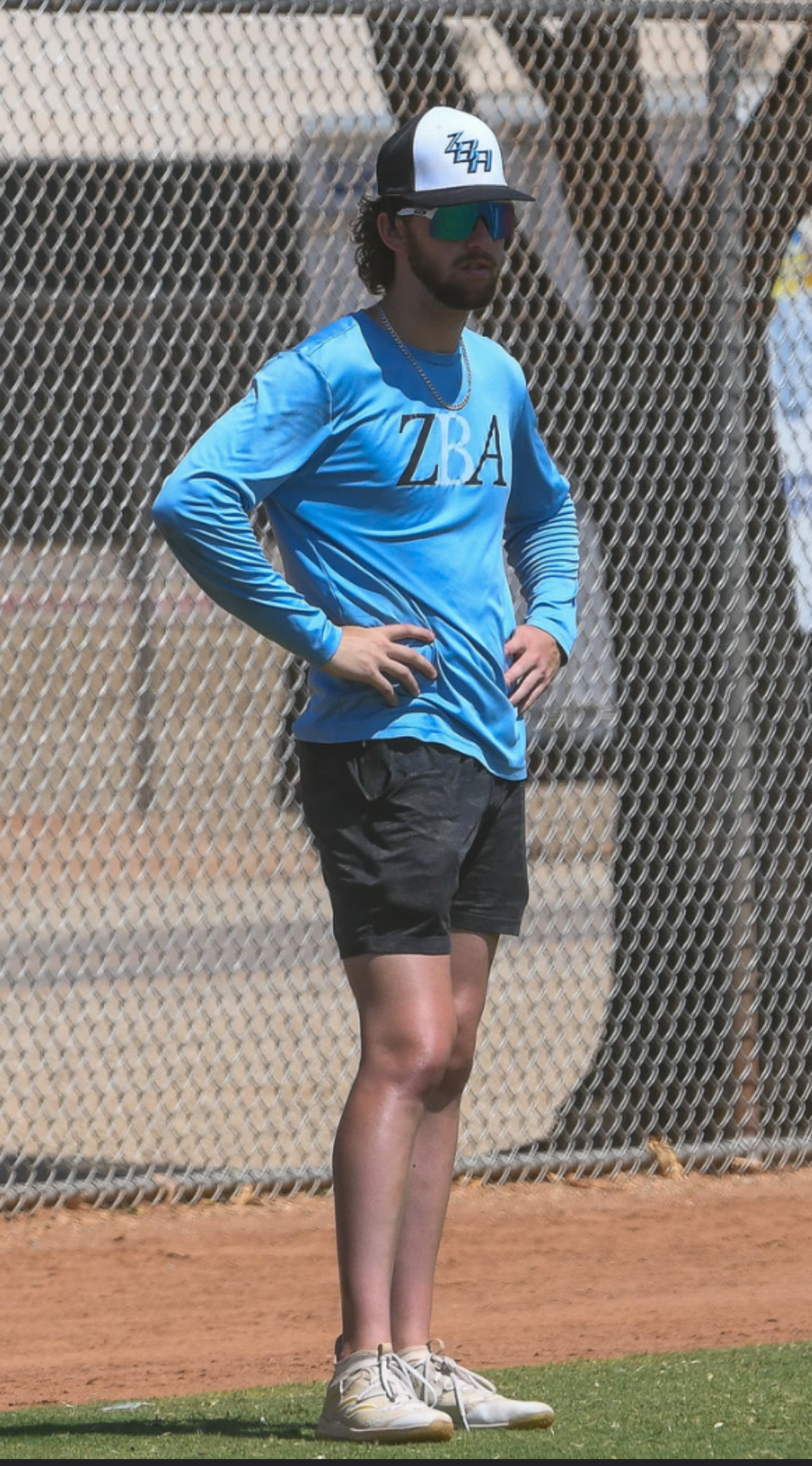 A man standing with hands on hips on a tennis court, wearing a blue long sleeve shirt, black shorts, white sneakers, a black and white cap, sunglasses, and a chain necklace, with a chain-link fence in the background.