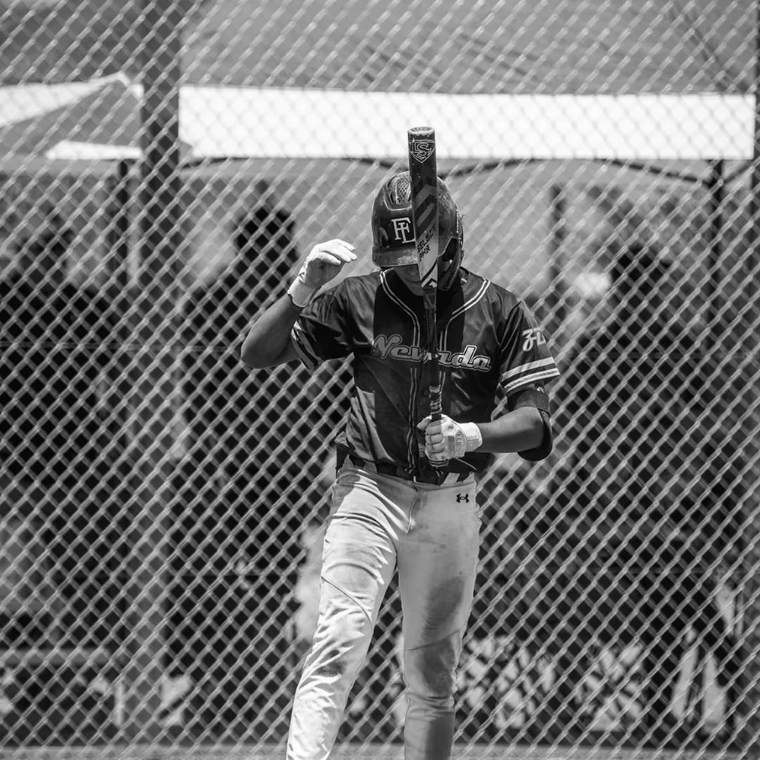 A baseball player standing behind a chain-link fence, holding a bat over his shoulder, wearing a helmet and uniform with the word 'Nevada'.