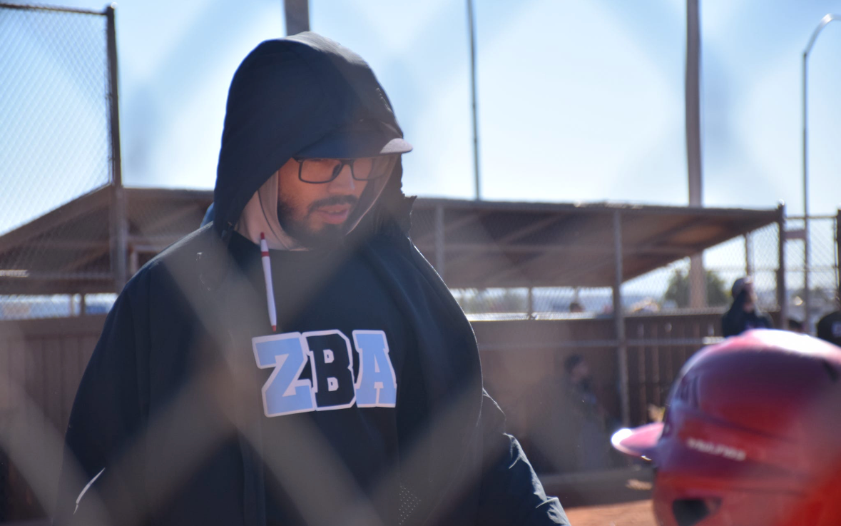 A man wearing glasses, a hoodie with the letters ZBA, and a cap under the hoodie, stands near a baseball field with a fence in the foreground and a clear sky in the background.