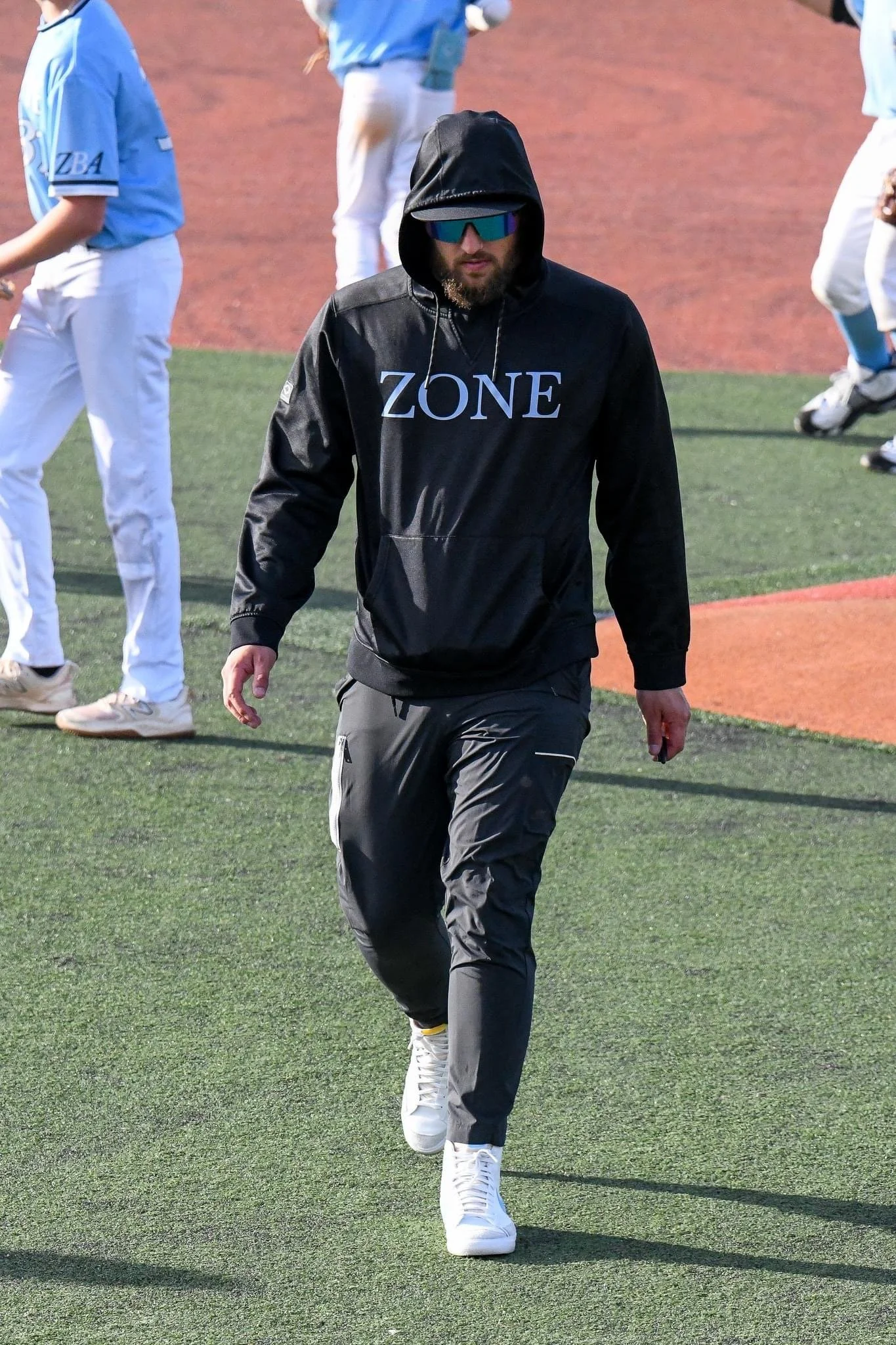 A person in a black hoodie with the word 'ZONE' on it, gray pants, white sneakers, sunglasses, walking on a baseball field with other players in the background.
