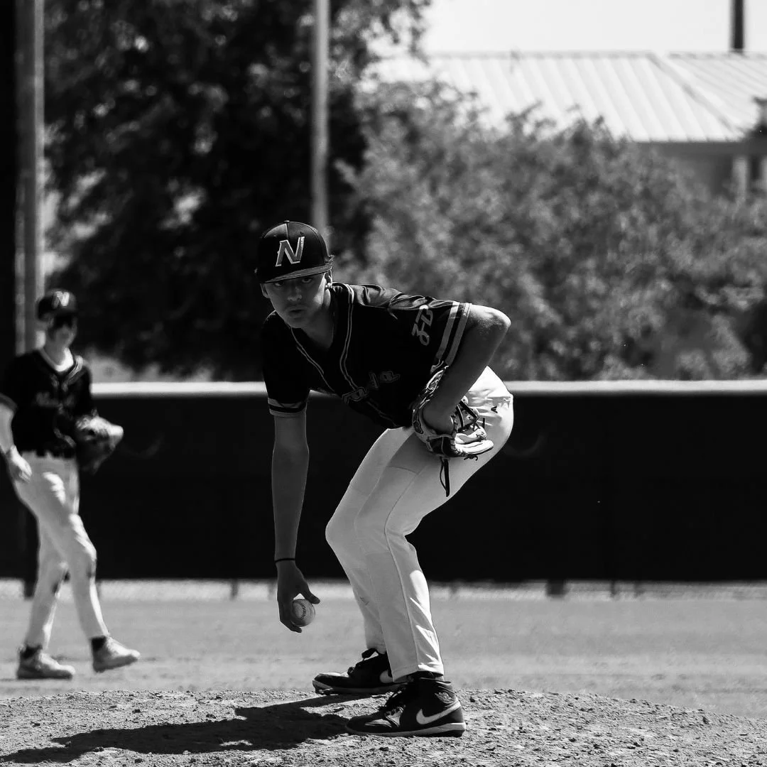 Black and white photo of a young baseball pitcher on the mound holding a baseball, with a teammate in the background on the field.