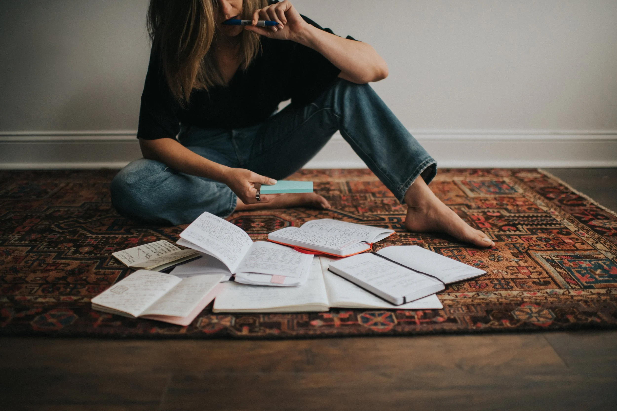 A woman sitting on a patterned rug on the floor surrounded by open notebooks and papers, holding a blue notepad and a pen, with additional notebooks and papers spread out around her.