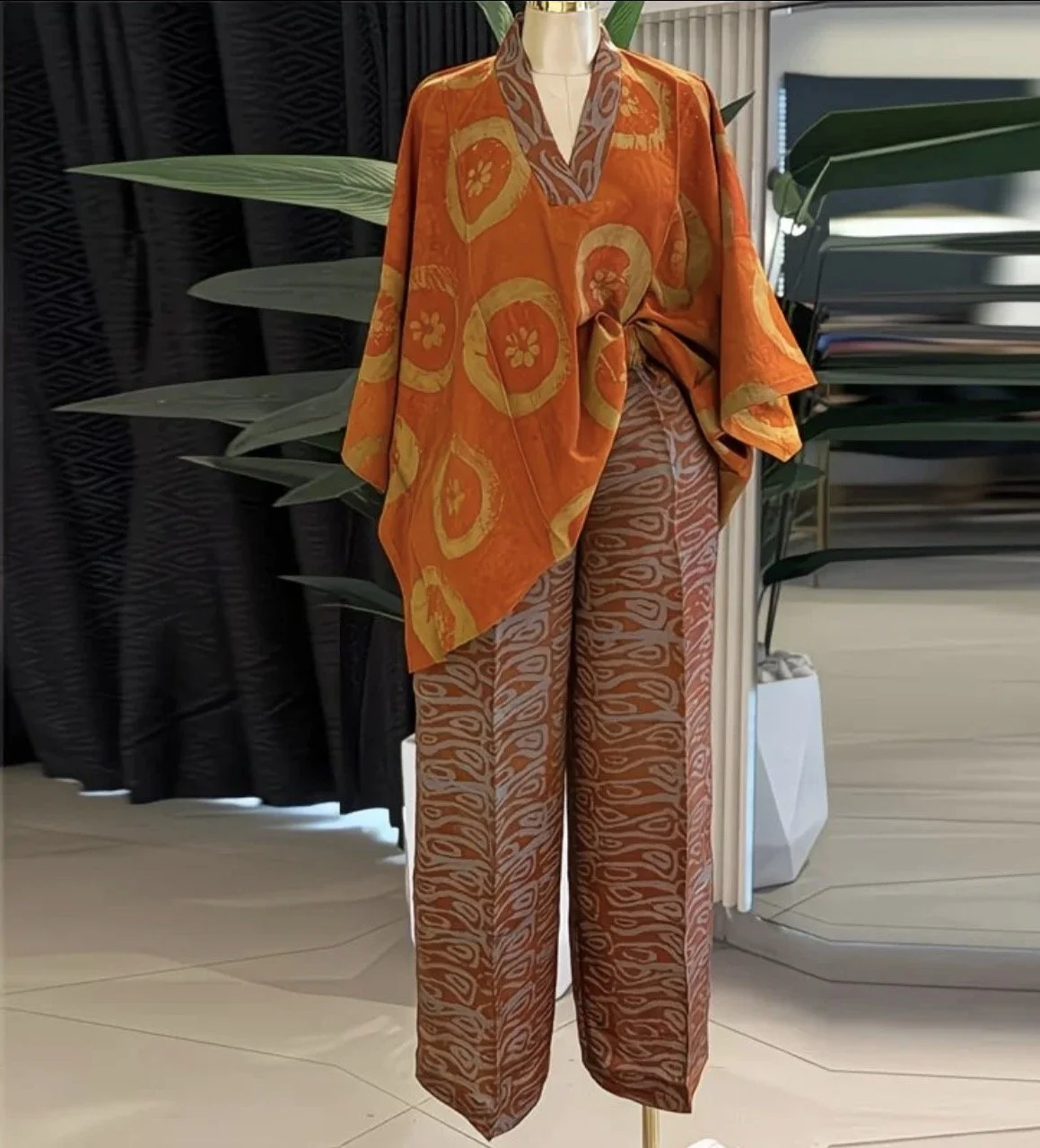 Mannequin dressed in an orange patterned kimono-style top with leaf and floral motifs, paired with wide-leg pants featuring an abstract animal print pattern, set against a black curtain and green plant background.