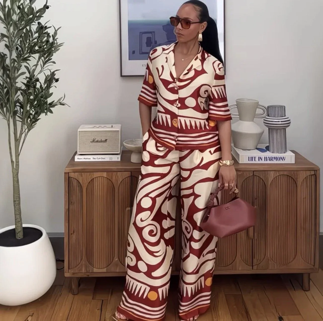 Woman wearing a matching abstract patterned shirt and pants in beige and dark red, holding a pink handbag, standing in front of a wooden cabinet with decorative vases and books, wearing sunglasses. cheekychk