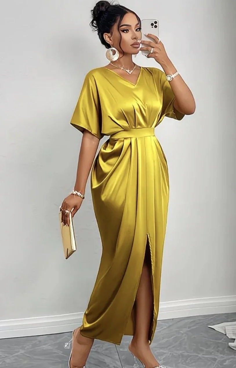 A woman taking a mirror selfie while wearing a shiny yellow satin dress with a slit, accessorized with large earrings, a necklace, a watch, and holding a clutch.