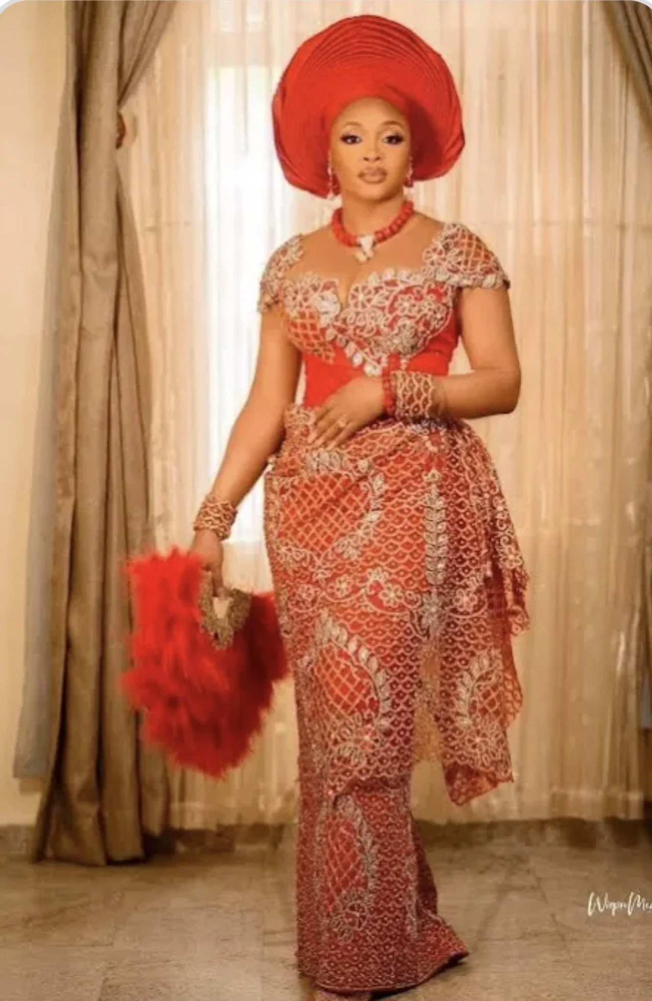 A woman dressed in traditional Nigerian attire, including a red gele (head wrap), a matching lace blouse, and a long patterned skirt, standing indoors in front of curtains. cheekychk
