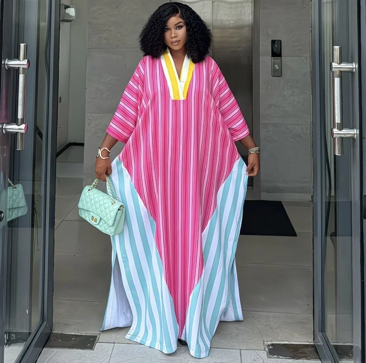 Woman in colorful striped kaftan holding a mint green quilted purse, standing in an entrance doorway with glass doors. cheekychk