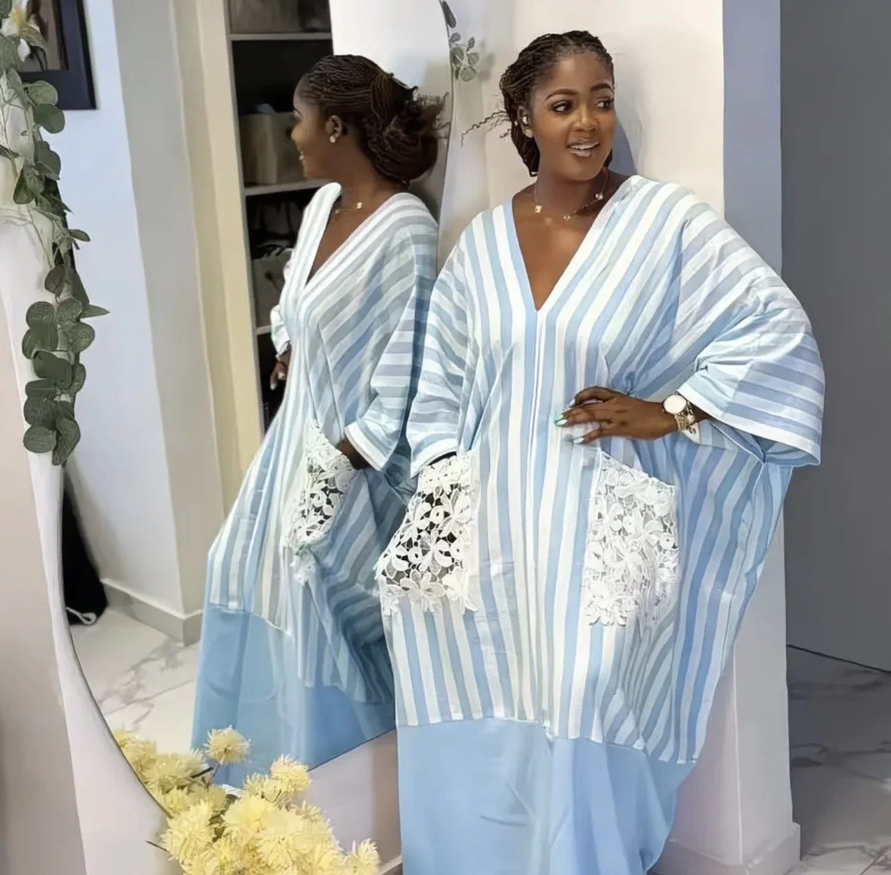 A woman standing in front of a mirror, wearing a blue and white striped dress with lace pockets, smiling and posing with one hand on her hip. Her reflection shows her profile with neatly styled hair, accessories, and jewelry, in a modern room with a 