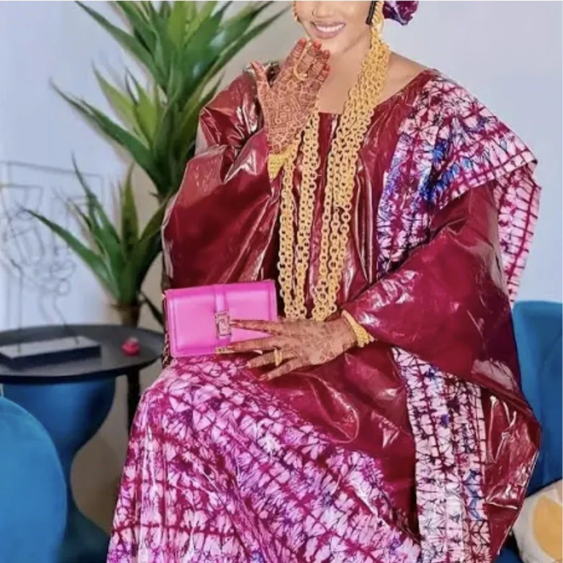 A woman wearing a traditional deep red and pink tie-dye dress with gold jewelry, holding a pink clutch, sitting on a blue chair near greenery. cheekychk