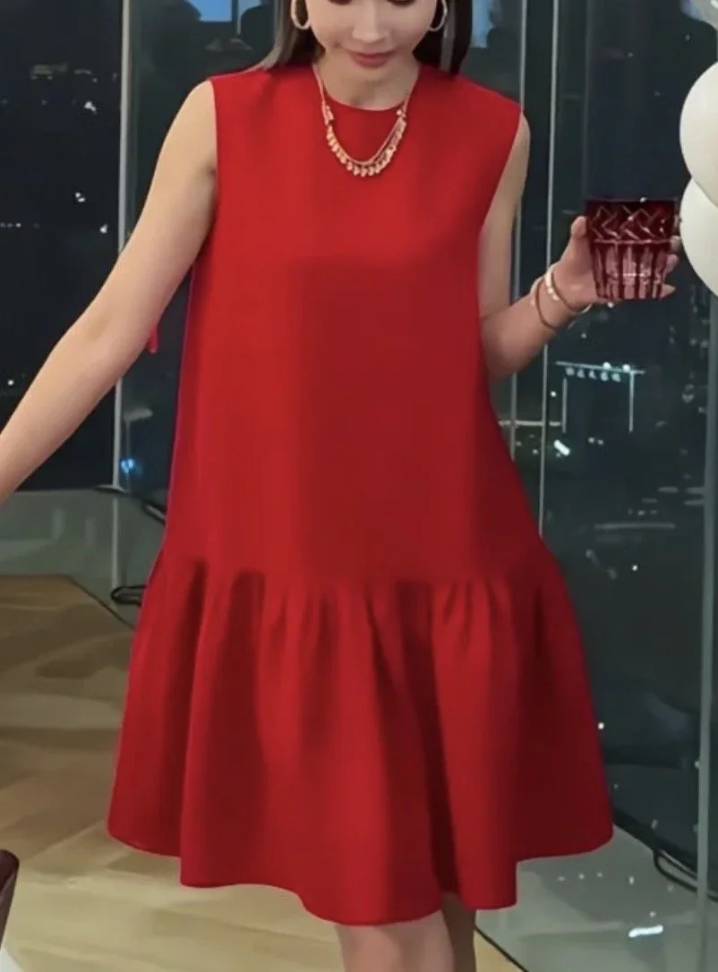 A woman in a sleeveless red dress holding a chocolate cupcake, wearing gold jewelry, in front of a cityscape at night.