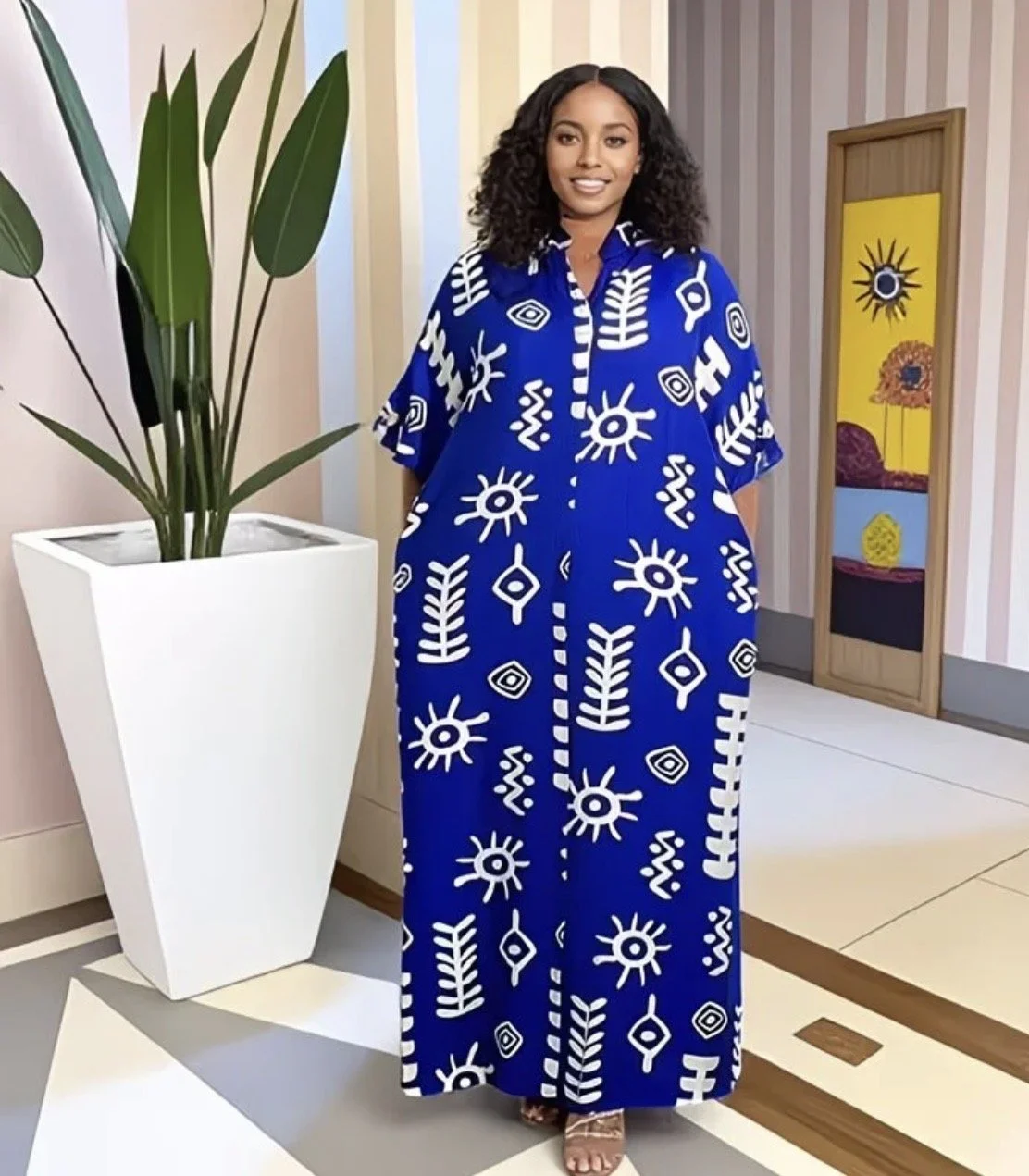 A woman wearing a long, blue dress with white tribal patterns stands indoors beside a tall potted plant with large green leaves, in front of colorful, striped wallpaper and a decorative door.