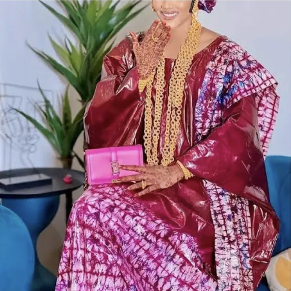 A woman dressed in traditional African attire with jewelry, sitting on a blue chair with a pink purse in her lap, and a green plant in the background. cheekychk