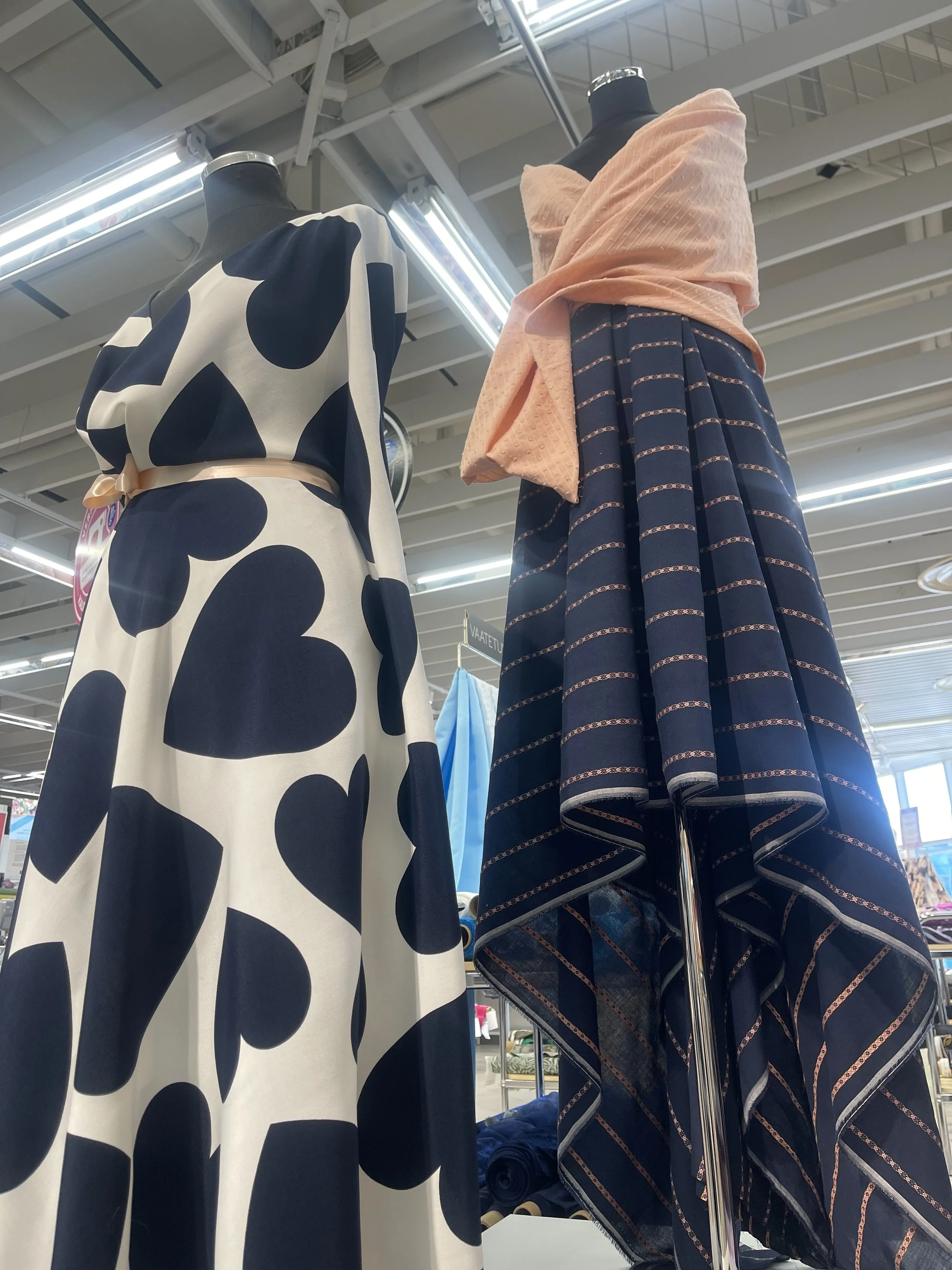Two mannequins displaying dresses inside a retail store with bright overhead lighting. The dress on the left has a white background with large black polka dots, and the dress on the right features a navy blue fabric with thin, pink and orange horizon