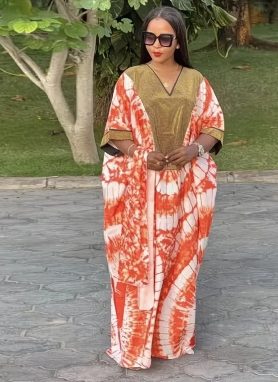 A woman standing outdoors on a paved surface, wearing a long, colorful, floral dress with orange, white, and pink patterns, black sunglasses, and silver jewelry, with trees and greenery in the background. cheekychk