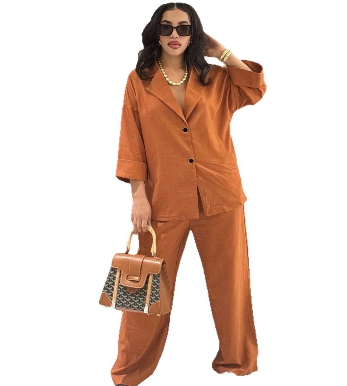 Woman wearing an orange blazer and matching wide-leg pants, black sunglasses, gold jewelry, holding a patterned handbag, posing with one hand near her hair.