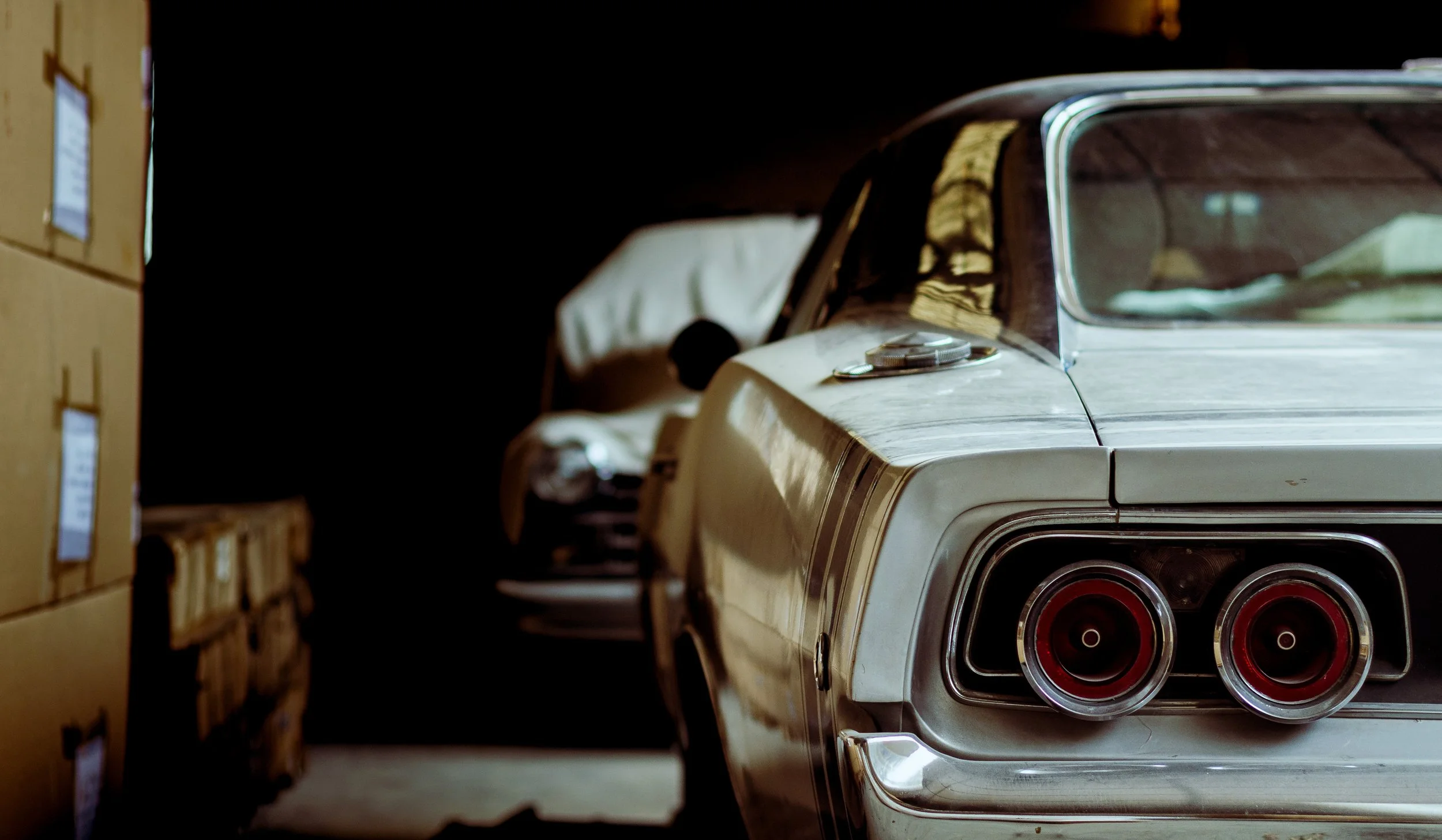 Close-up of the rear end of a classic white car with three round tail lights, parked in a dimly lit storage area with cardboard boxes on the left side.
