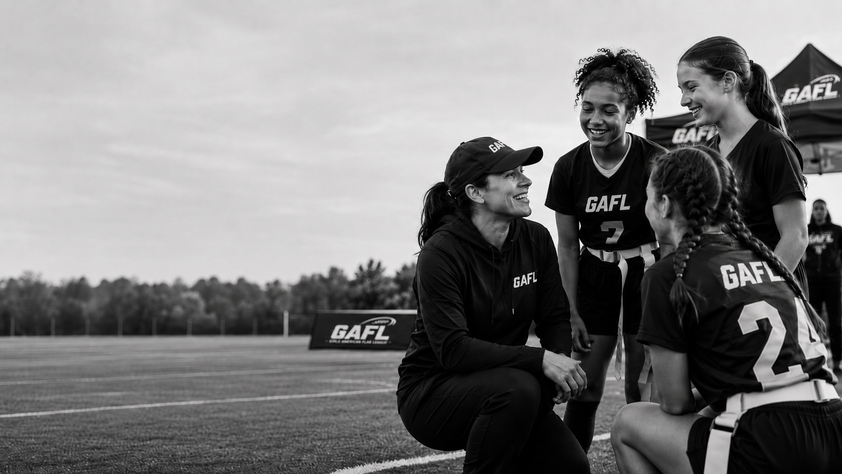 A group of female athletes and their coach having a discussion on a sports field. They are wearing uniforms with 'GAFL' printed on them, some are standing and others are kneeling, and they are smiling and engaging with each other.