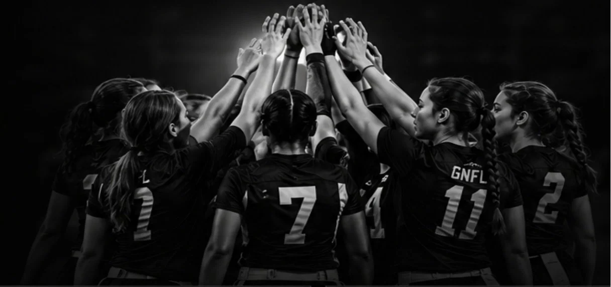 A group of female flag football players in black jerseys with numbers and letters on their backs, huddled together with their hands stacked in the center, preparing for a game or celebration.